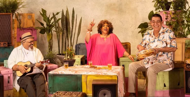 Fãs ganham nova chance: Alcione, Jorge Aragão e Zeca Pagodinho anunciam data extra 1 Grupo musical com instrumentos sentados em mesa rodeada de plantas e bebidas durante show temático.