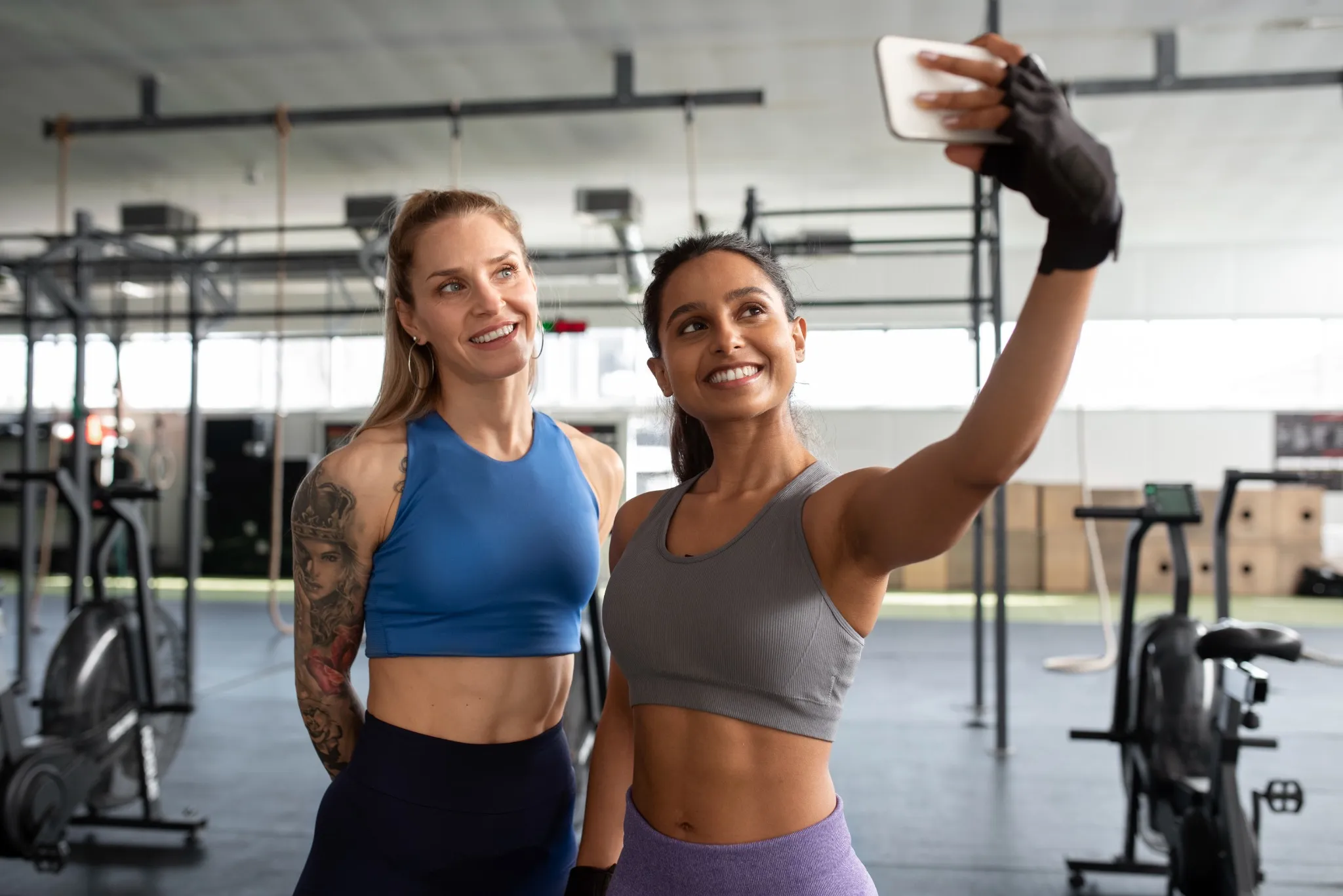 Segundo a ciência, pessoas que postam muitas fotos de exercícios na academia podem estar escondendo este sentimento específico 2 Duas mulheres sorrindo e tirando uma selfie na academia, exemplificando a relação entre postagens de treino e busca por validação.