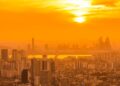 Vista aérea de uma metrópole densamente povoada sob um céu intensamente alaranjado e enevoado devido a uma forte onda de calor, com o sol brilhando entre brumas.