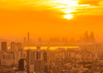 Vista aérea de uma metrópole densamente povoada sob um céu intensamente alaranjado e enevoado devido a uma forte onda de calor, com o sol brilhando entre brumas.