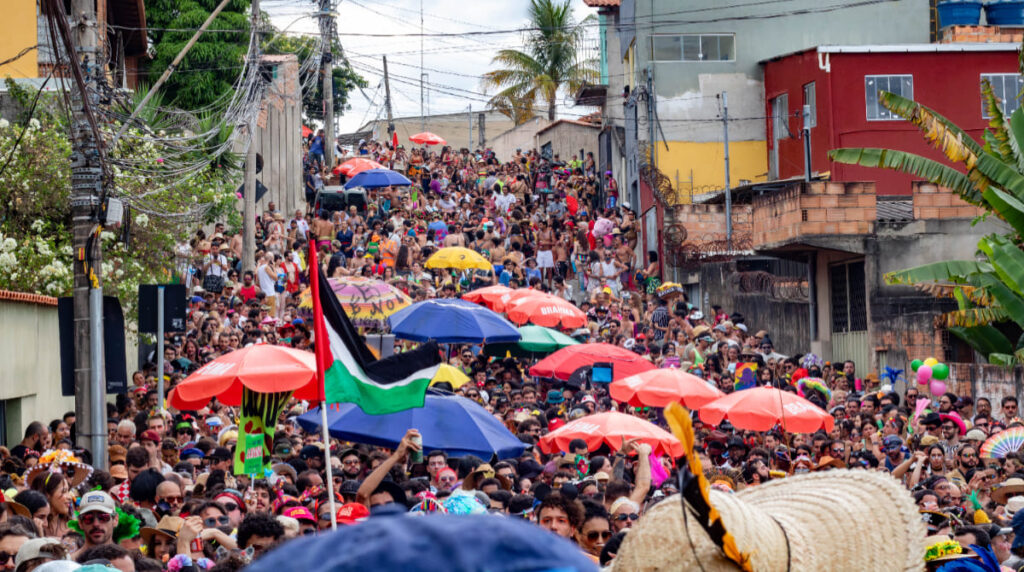 Vai chover no Carnaval? Veja a previsão para as principais capitais da folia em 2026 2 Pessoas fantasiadas segurando guarda-chuvas coloridos reunidas em bloco de Carnaval na rua