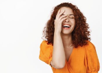 Jovem de cabelos cacheados e blusa laranja rindo abertamente enquanto cobre parcialmente o rosto com a mão, sobre um fundo branco.