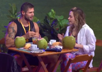 Participantes Jonas e Ana Paula sentados à mesa com drinks de coco durante conversa em clima de tensão e flerte durante festa no BBB 26