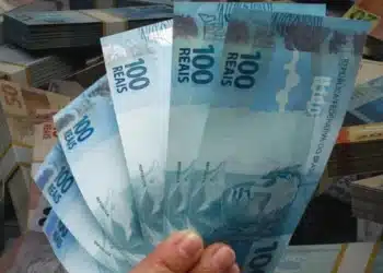 Mão segurando cédulas de real com outras notas ao fundo, representando o saque de até R$ 6.220 do FGTS para trabalhadores de cidades afetadas.