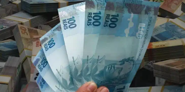 Mão segurando cédulas de real com outras notas ao fundo, representando o saque de até R$ 6.220 do FGTS para trabalhadores de cidades afetadas.
