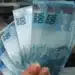 Mão segurando cédulas de real com outras notas ao fundo, representando o saque de até R$ 6.220 do FGTS para trabalhadores de cidades afetadas.