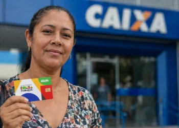 Beneficiária segura cartão do Bolsa Família em frente a uma agência da Caixa.