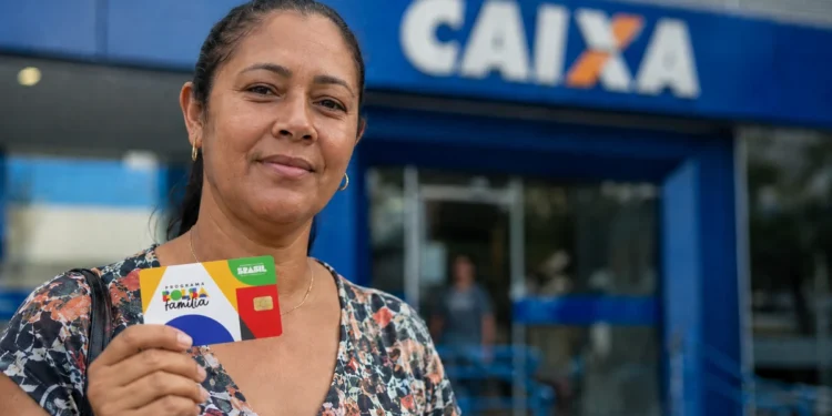 Beneficiária segura cartão do Bolsa Família em frente a uma agência da Caixa.