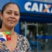 Beneficiária segura cartão do Bolsa Família em frente a uma agência da Caixa.
