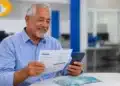 Aposentado consultando documento e celular em agência, representando o pagamento de atrasados do INSS.