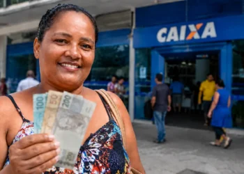 Mulher segura notas de reais em frente a uma agência da Caixa, representando o pagamento do Bolsa Família nesta sexta-feira (21 de agosto).
