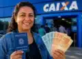 Mulher segura carteira de trabalho e notas de real em frente a uma agência da Caixa, representando a solicitação do Saque Calamidade do FGTS.