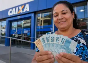 Mulher segura cédulas de real em frente a uma agência da Caixa, representando a solicitação do Saque Calamidade do FGTS até 29 de julho de 2026.