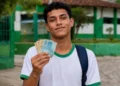 Estudante segura cédulas de dinheiro em frente à escola, representando o calendário de pagamentos do Pé-de-Meia de setembro.
