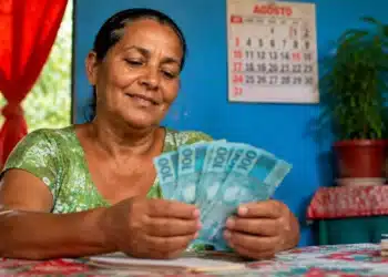 Beneficiária do programa Mães de Pernambuco segura notas de dinheiro em casa, representando o pagamento de R$ 300 em agosto.