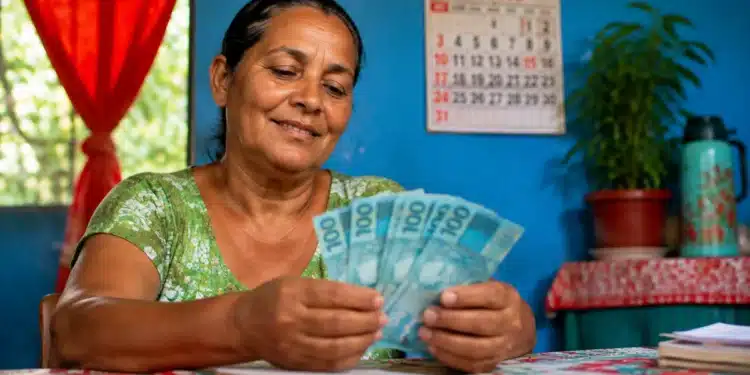 Beneficiária do programa Mães de Pernambuco segura notas de dinheiro em casa, representando o pagamento de R$ 300 em agosto.