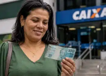 Beneficiária do Bolsa Família segura nota de R$ 100 em frente a uma agência da Caixa.