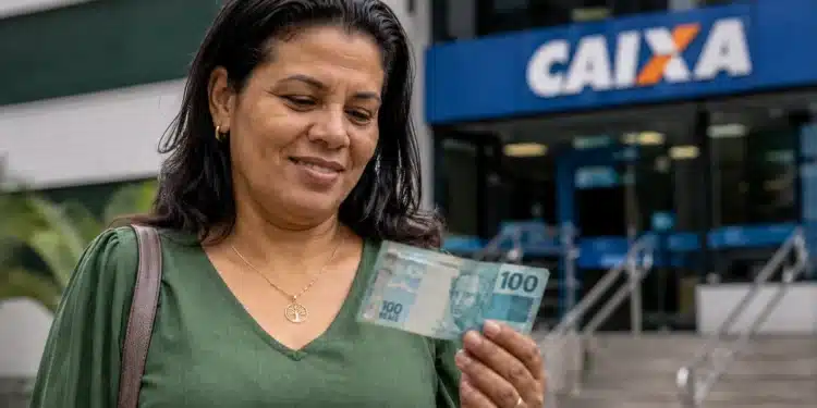 Beneficiária do Bolsa Família segura nota de R$ 100 em frente a uma agência da Caixa.