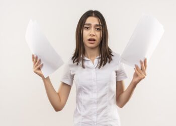 young girl in white shirt holding blank pages looking at camera confused standing over white background