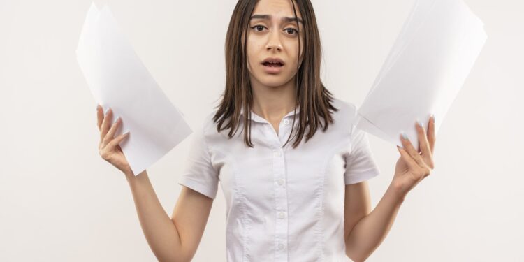 young girl in white shirt holding blank pages looking at camera confused standing over white background