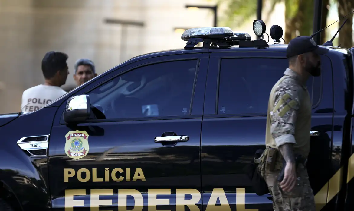 Viatura da Polícia Federal com agentes ao fundo e policial tático ao lado.