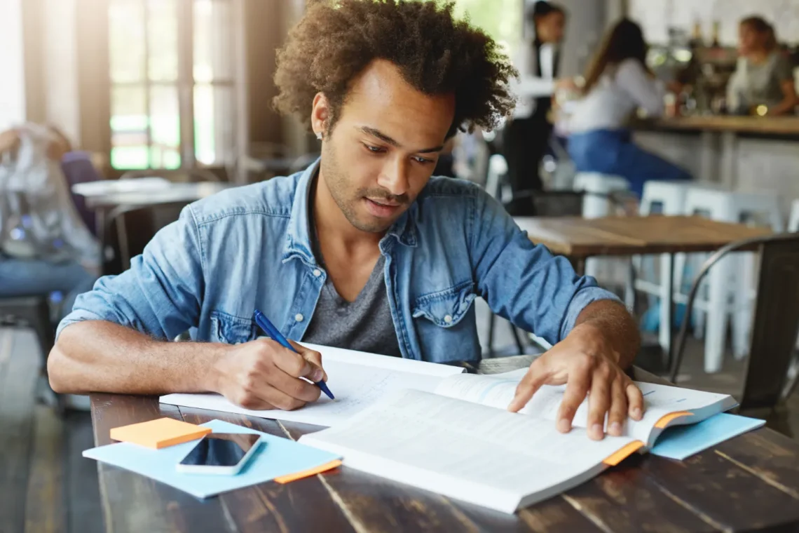 Homem com cabelo cacheado estudando em uma mesa, com livros e material de escritório, focado na preparação para o concurso