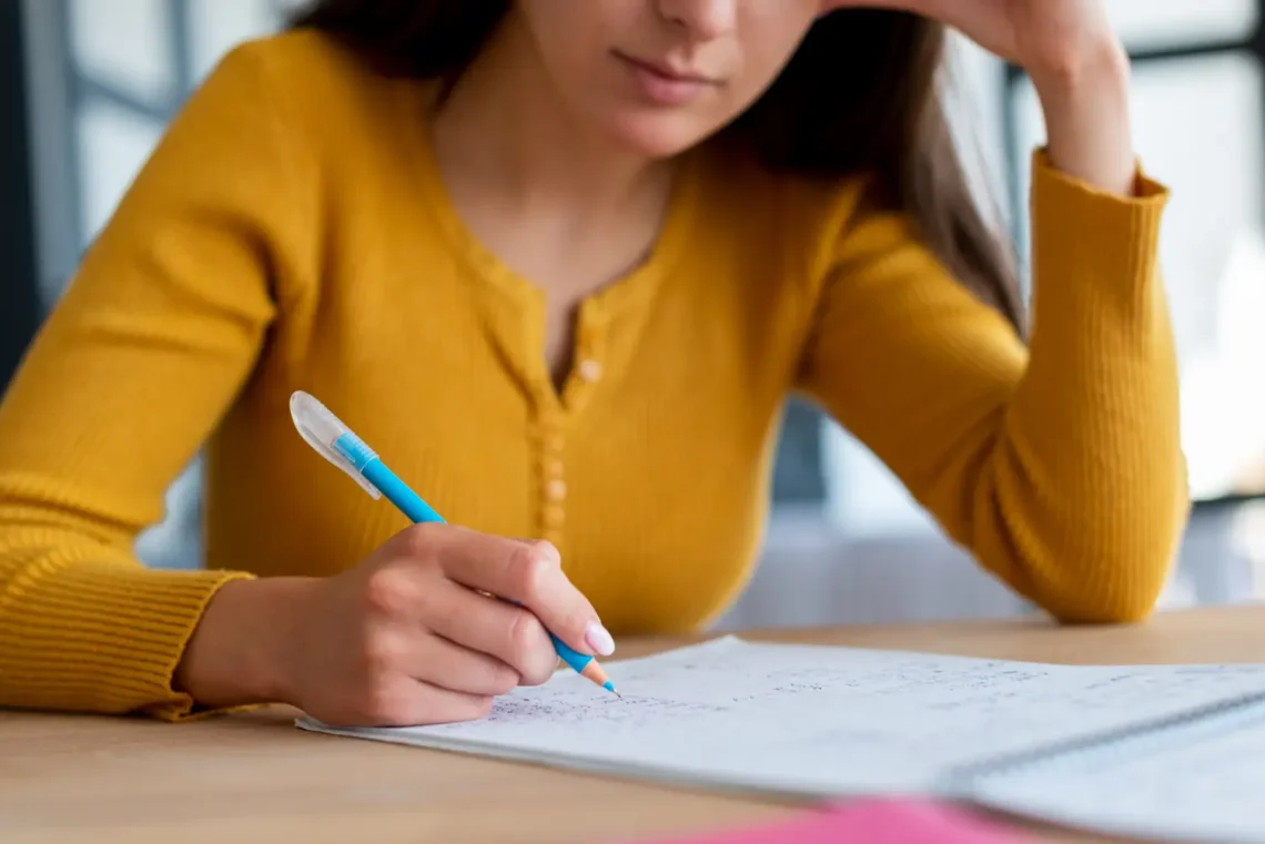 Mulher estudando com uma caneta azul, fazendo anotações em uma folha de papel enquanto se concentra no material de estudo