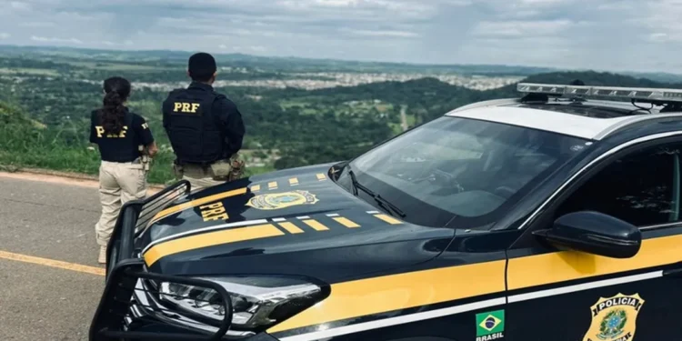 Dois agentes da PRF de costas observando uma rodovia, ao lado de uma viatura oficial