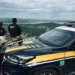 Dois agentes da PRF de costas observando uma rodovia, ao lado de uma viatura oficial