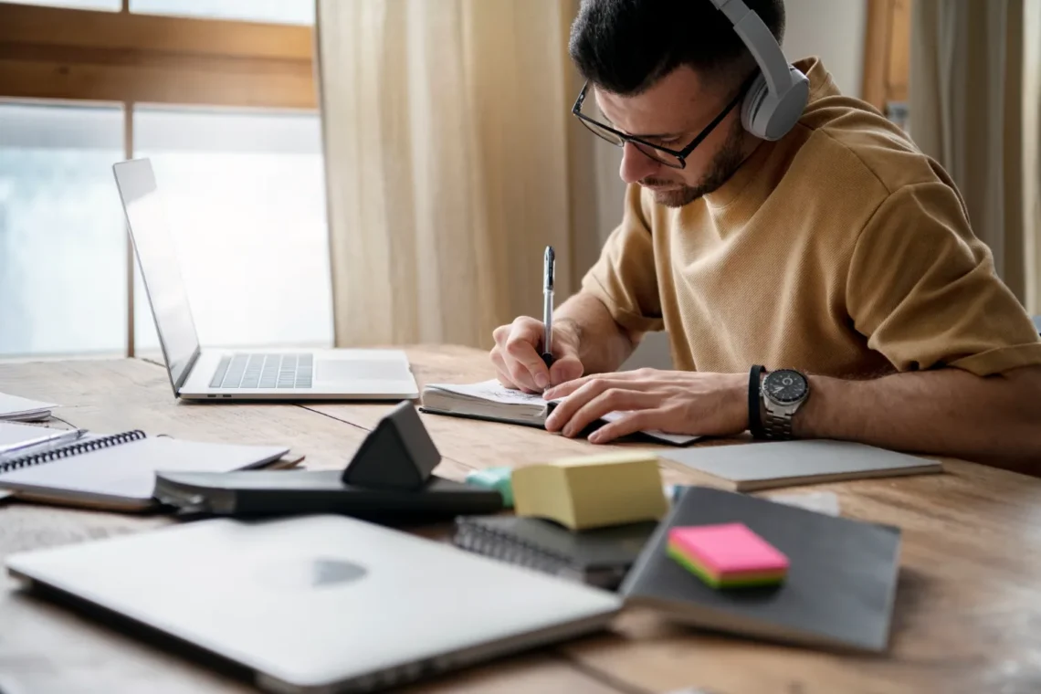 Homem estudando com fones de ouvido, escrevendo em um caderno ao lado de um notebook