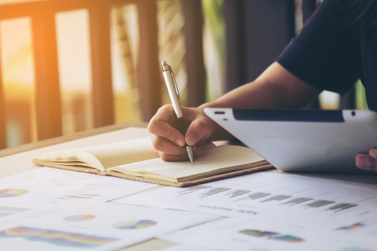 Pessoa anotando informações em um caderno enquanto utiliza um tablet, com gráficos e documentos na mesa