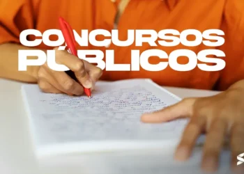 Pessoa escrevendo redação com caneta vermelha em folha de papel, representando concursos públicos.