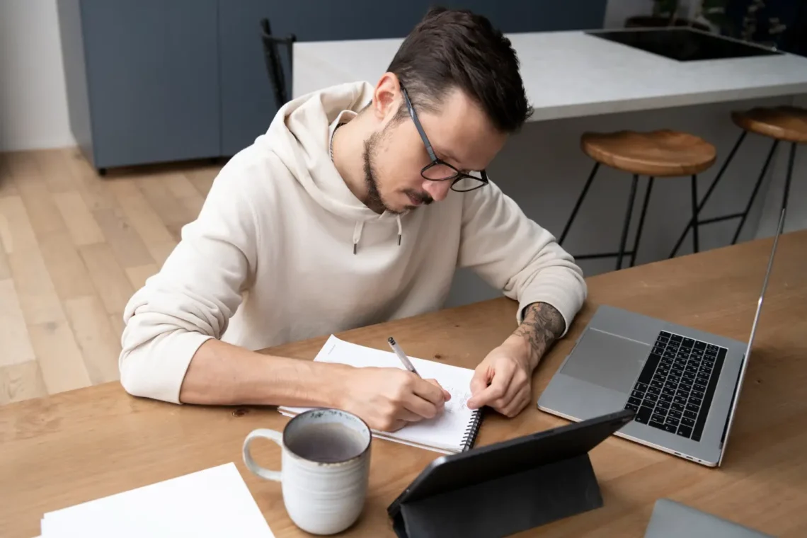 Homem estudando com caderno e computador, focado na preparação para o concurso de Agente Administrativo da Polícia Federal