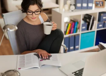 Pessoa trabalhando e estudando ao mesmo tempo, lendo um livro e tomando café, representando o equilíbrio entre estudo e trabalho.