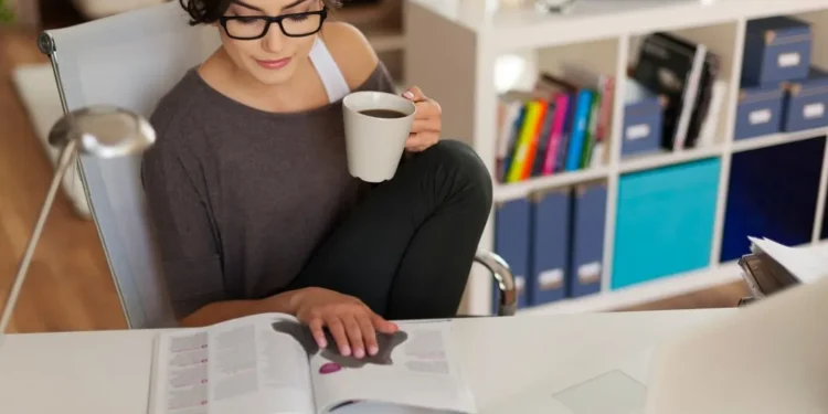 Pessoa trabalhando e estudando ao mesmo tempo, lendo um livro e tomando café, representando o equilíbrio entre estudo e trabalho.