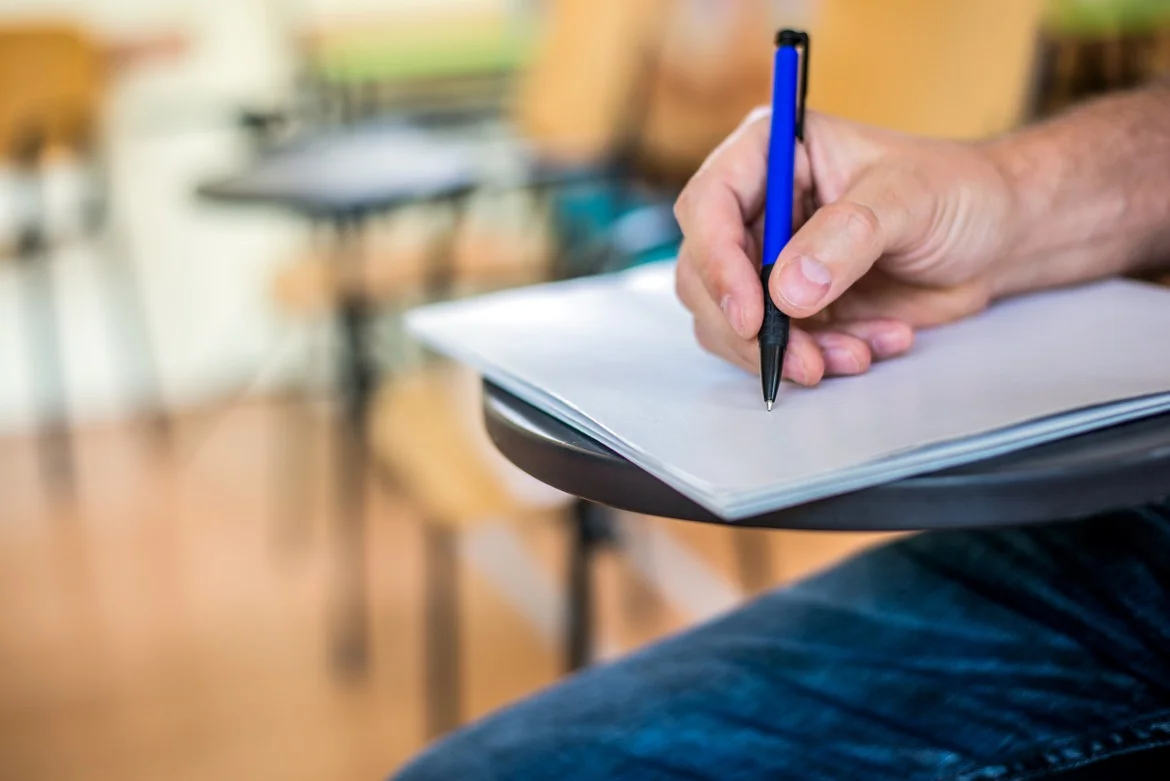 Pessoa escrevendo em um caderno sobre uma mesa de sala de aula, representando inscrição em concurso público.