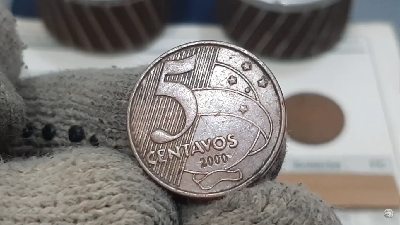 Moeda rara de 5 centavos do ano 2000 sendo segurada com luvas, destacando detalhes e relevos.