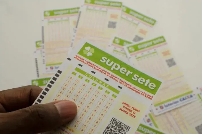 Mão segurando um bilhete da loteria Super Sete, com vários outros bilhetes ao fundo