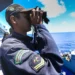Militar da Marinha do Brasil observando o mar com binóculos em uma embarcação.