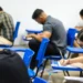 Grupo de pessoas realizando provas em sala de aula, com foco em um estudante escrevendo.