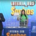 Apresentadores do sorteio da Loteria dos Sonhos, com um globo de números e uma casa transparente