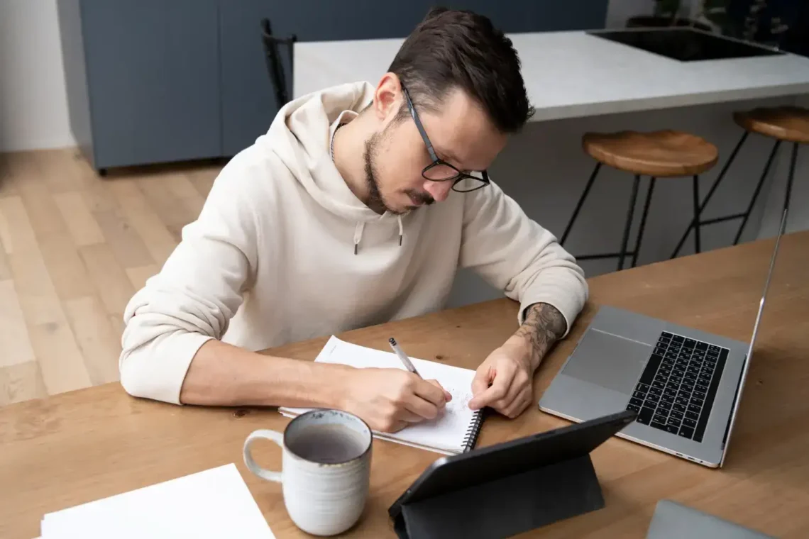 Homem estudando com caderno, tablet e laptop em uma mesa de madeira, preparando-se para um concurso público