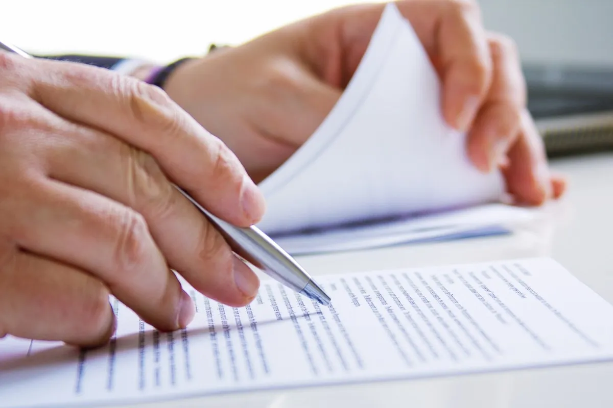 Mão segurando caneta e revisando documentos, representando o processo de inscrição no concurso público.