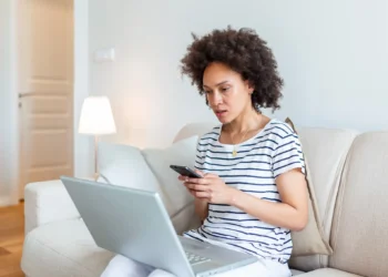 Mulher sentada no sofá, usando laptop e celular enquanto trabalha no Currículo Lattes.