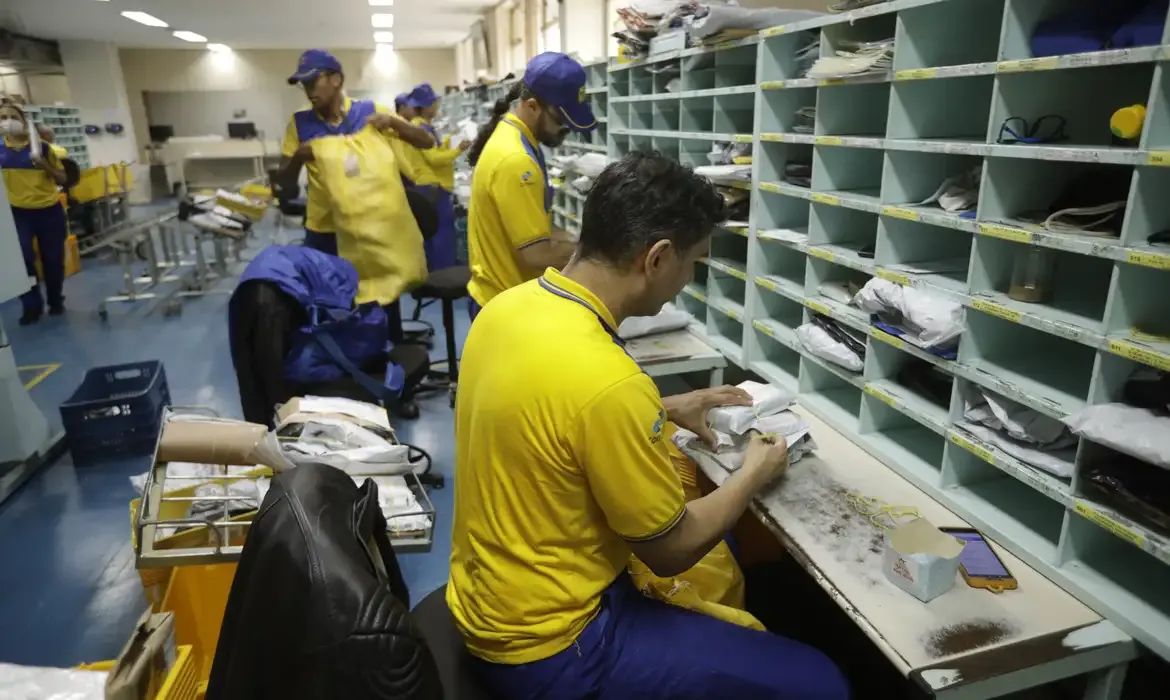 Trabalho em centro de distribuição postal Funcionários organizando pacotes em um centro de distribuição postal dos Correios, com vários compartimentos de armazenamento.