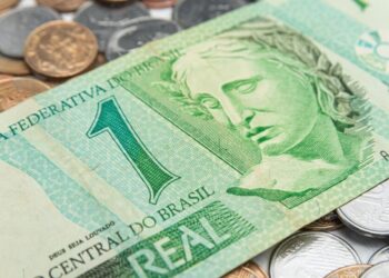 Imagem de uma cédula de 1 real em primeiro plano, com moedas de diferentes valores ao fundo, ilustrando o tema das cédulas do real que serão retiradas de circulação.