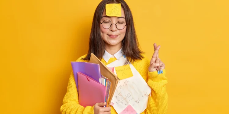 Estudante sorridente cruzando os dedos para boa sorte, com documentos e pastas.
