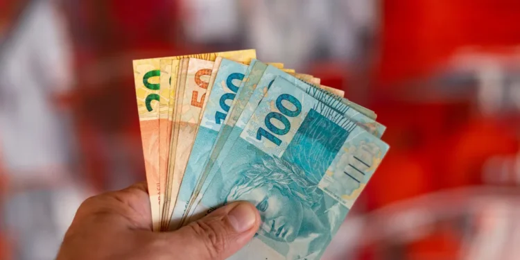 Notas de dinheiro de diferentes valores (R$ 20, R$ 50, R$ 100) sendo seguradas por uma mão, com fundo desfocado.