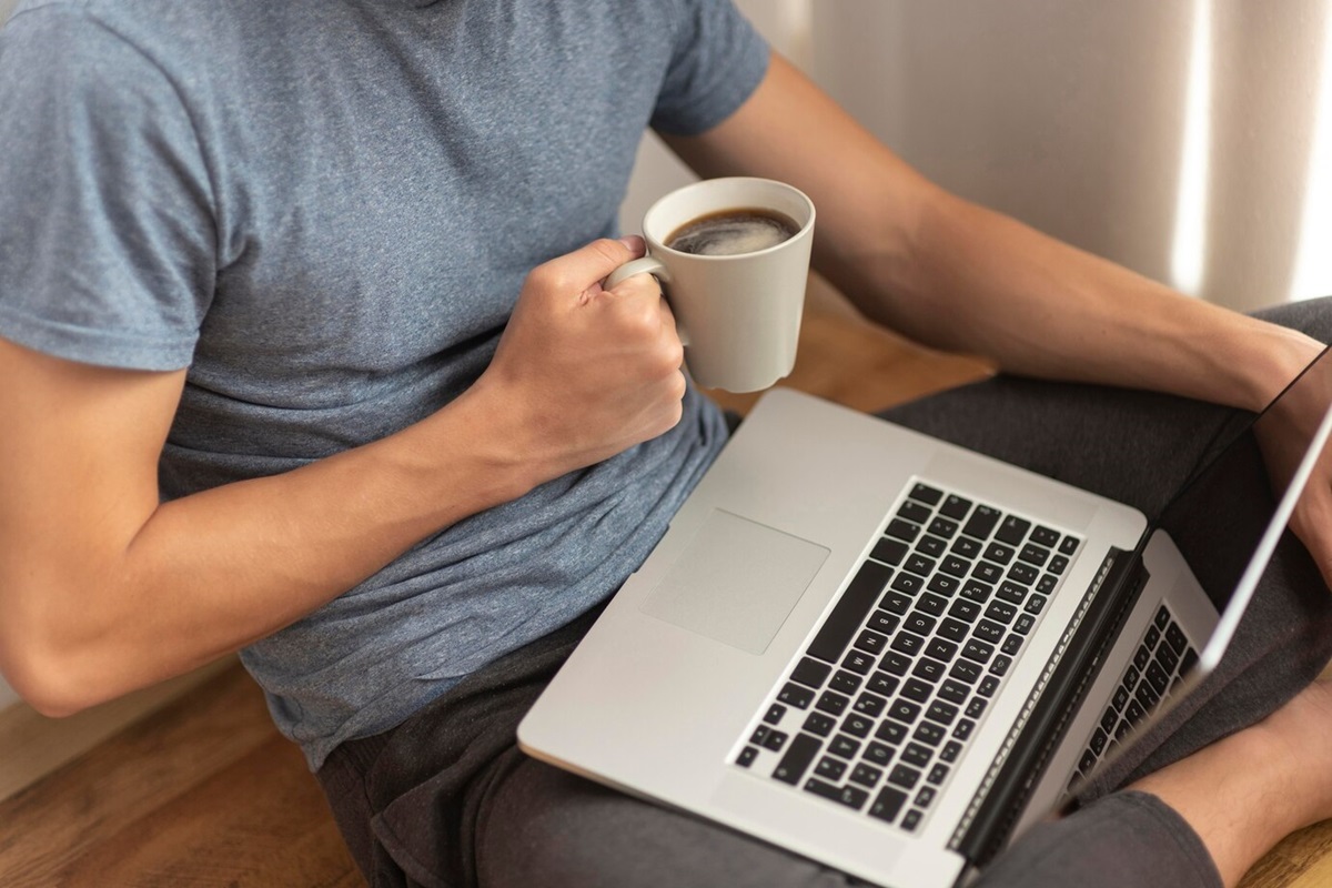 Confira as dicas para evitar que seu notebook danifique. Homem tomando café com seu notebook no colo.