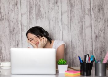 Mulher com fones de ouvido e expressão cansada usando notebook em mesa com itens de escritório